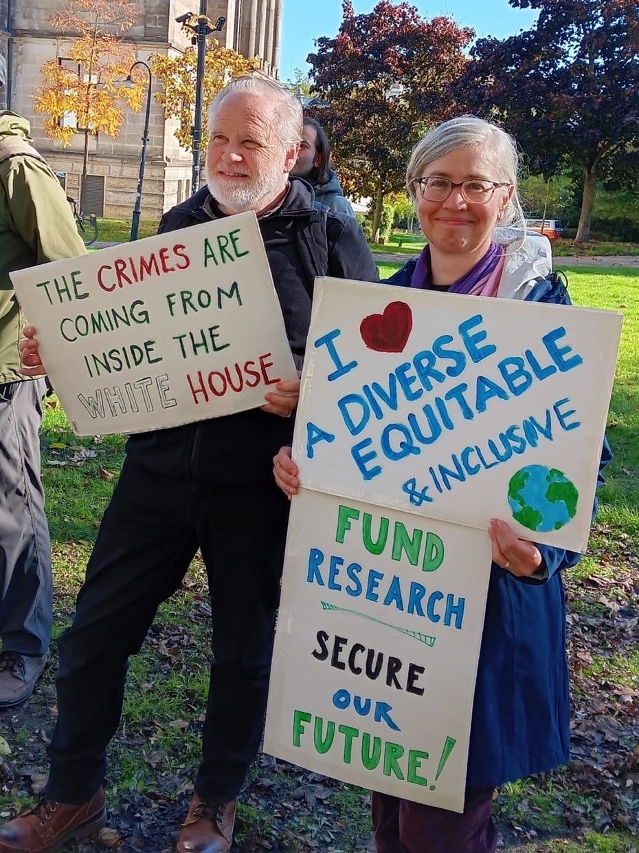 Two protesters holding protest signs about U.S. policies and democratic values in a public park. One sign reads 'The crimes are coming from inside the White House'. Another signs says 'Fund research - secure our future'. And the third sign says 'I love a diversive equitable & inclusive earth'